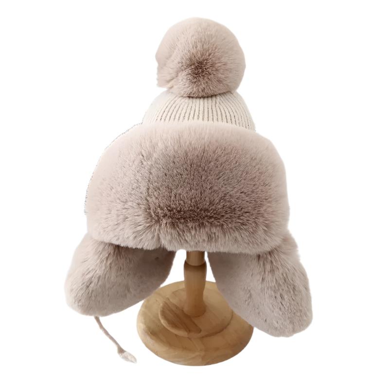Person iført beige unisex trapper lue med øreklaffer og pompong, perfekt for kalde dager utendørs.
